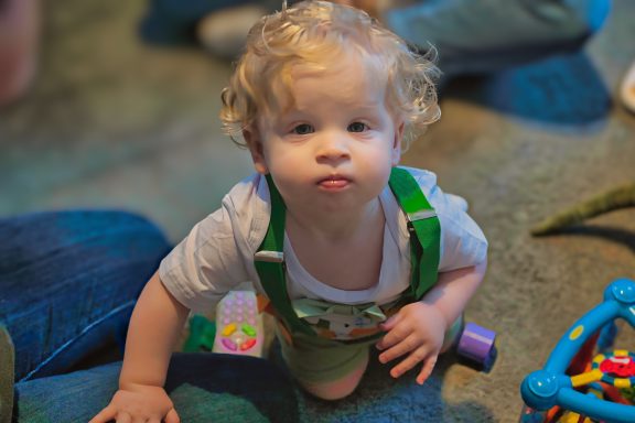Baby Posing Toddler with curly hair wearing green overalls, sitting on the floor with toys nearby.