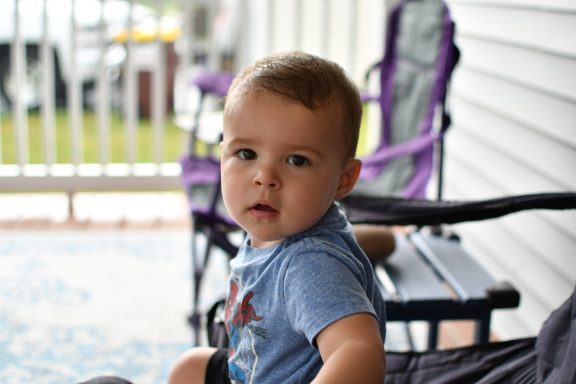 Child suprise pic Toddler sitting on a porch with chairs in the background, looking curiously at the camera.